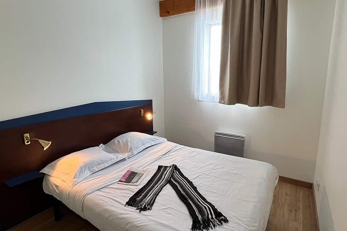 A neatly made bed with white linens, a striped blanket, and a pillow. There is a bedside table with a lamp and a small object on it. A window with curtains is visible, and a radiator is on the wall.