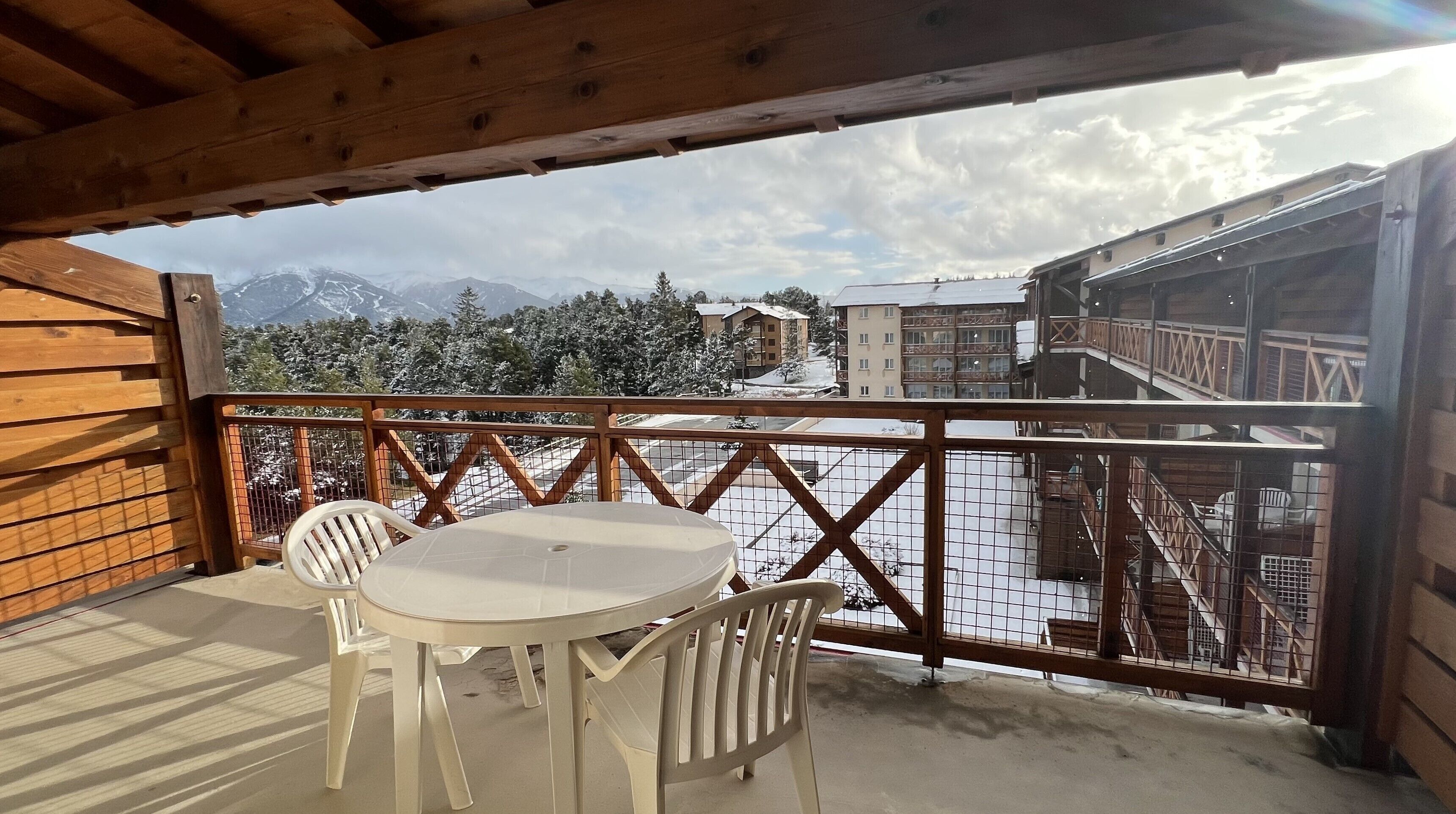 Un balcon en bois avec une table et des chaises, donnant sur un paysage enneigé parsemé de bâtiments et d’arbres.