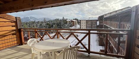 Un balcon en bois avec une table et des chaises, donnant sur un paysage enneigé parsemé de bâtiments et d’arbres.
