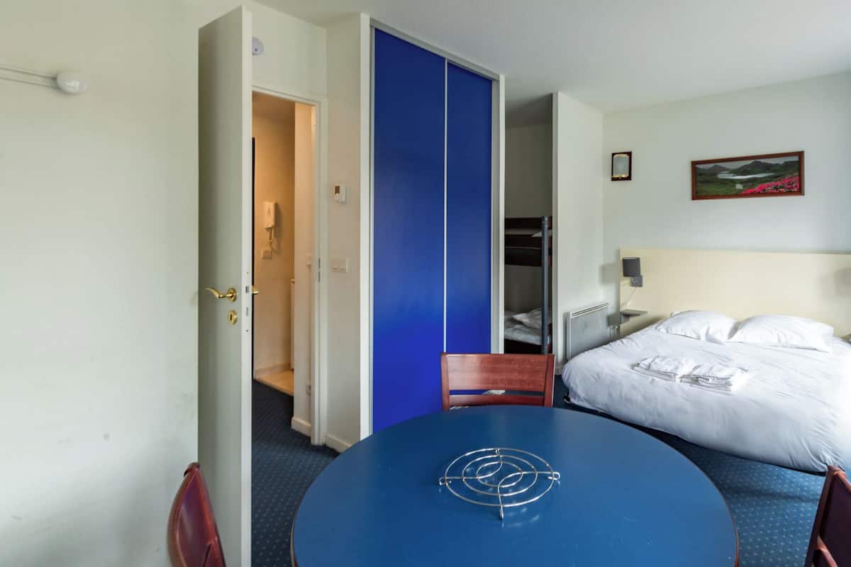 A hotel room with a bed, a desk, a chair, a blue table, a wardrobe, and a painting on the wall.