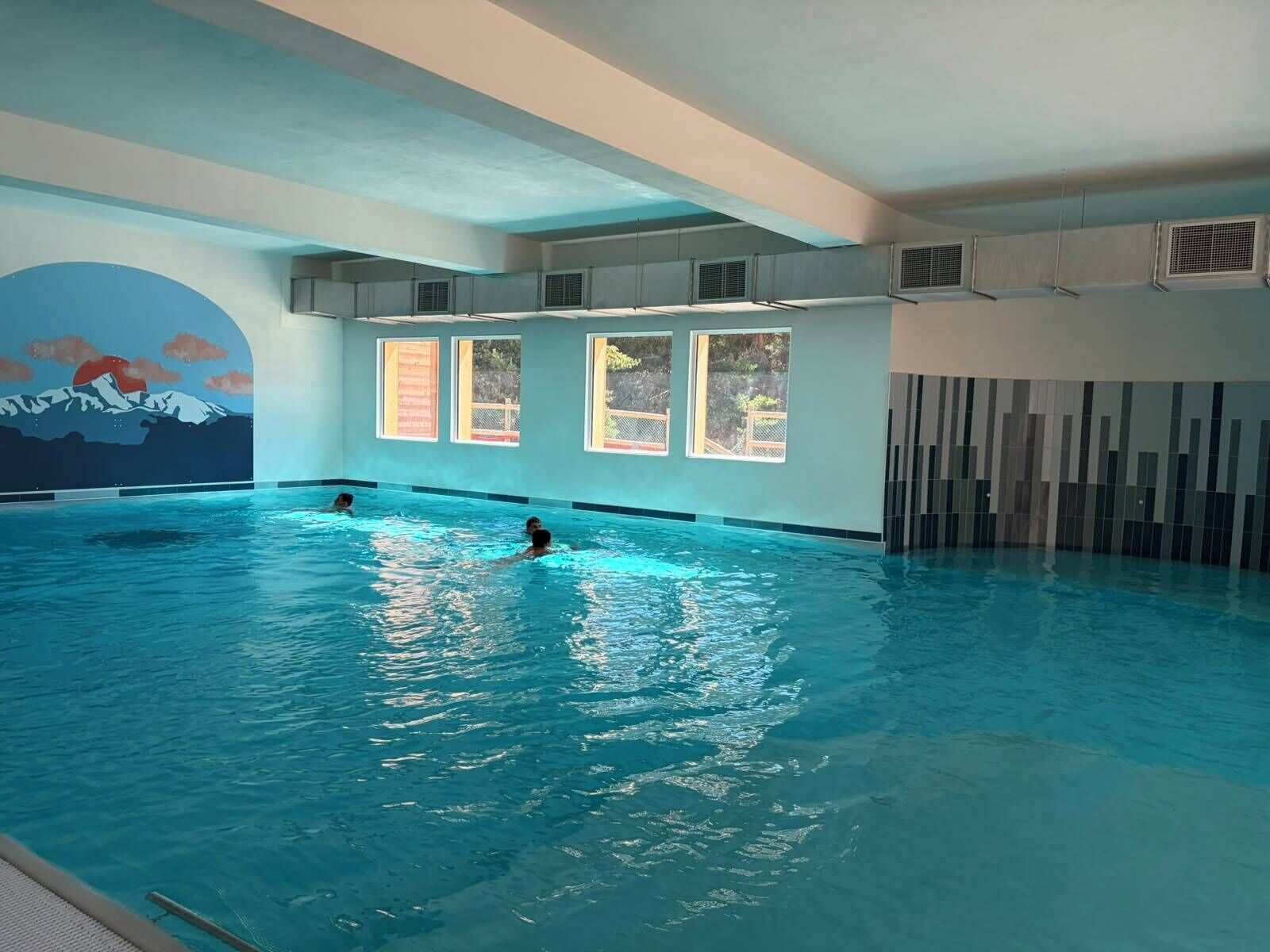 An indoor swimming pool with clear water, a mural on the wall, and windows allowing natural light.