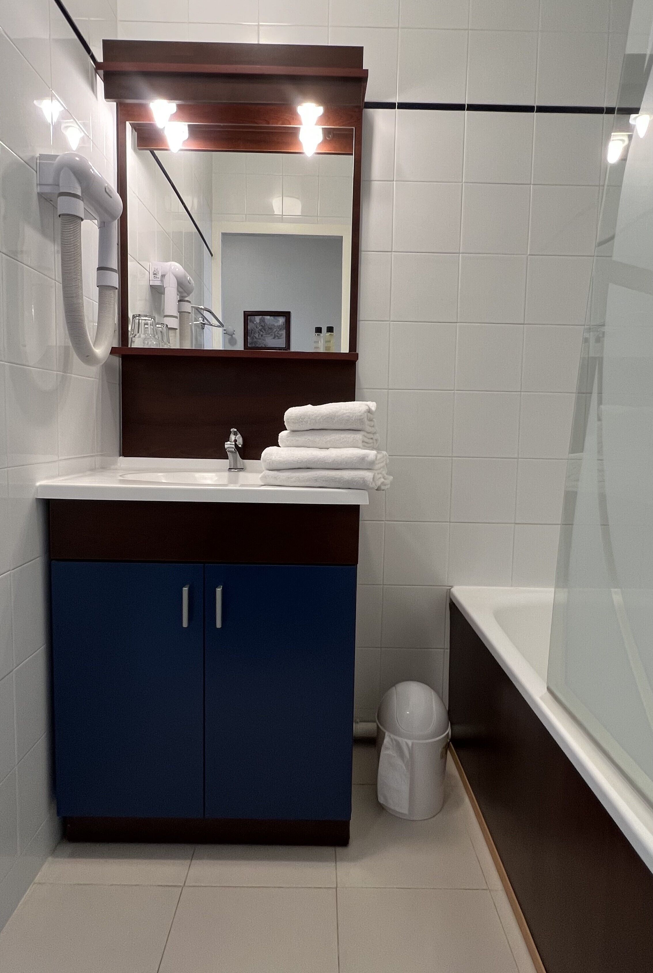 A modern bathroom with a white sink, blue cabinets, a mirror with lights, a hairdryer, and a glass shower enclosure.