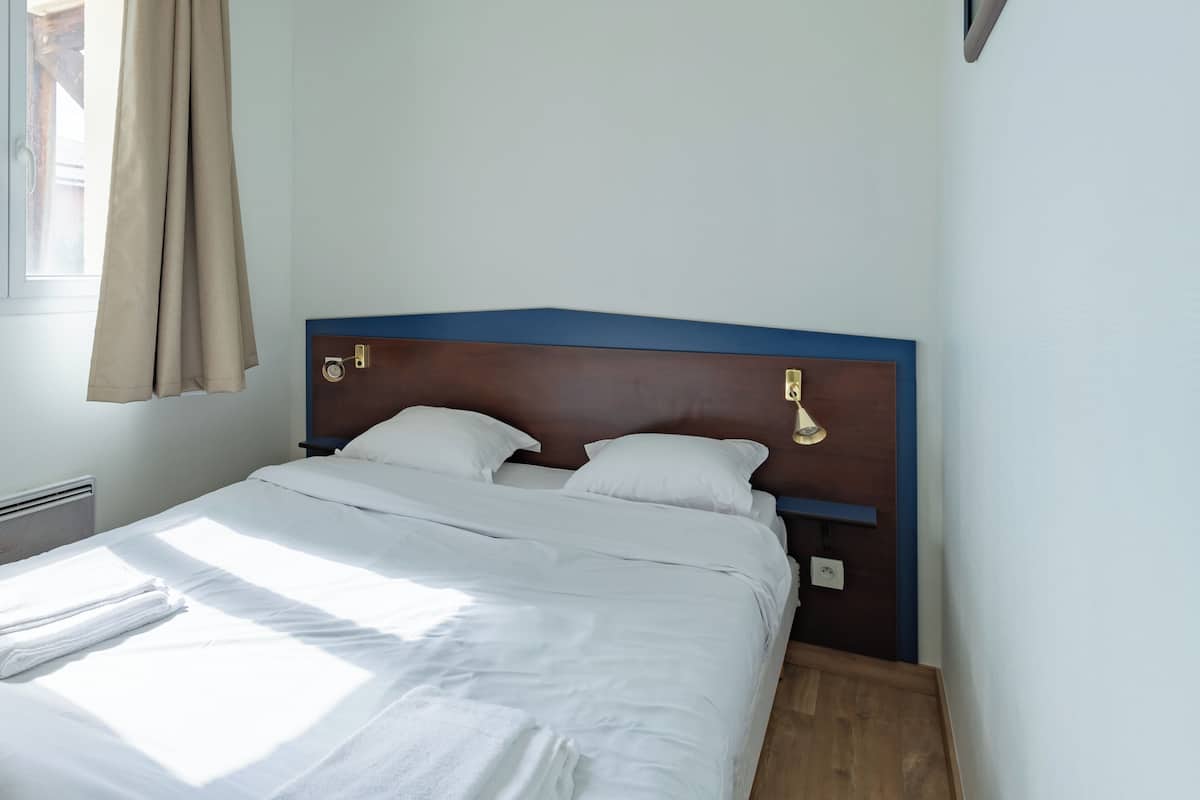A neatly made bed with white linens, a wooden headboard, and a wall-mounted light fixture.