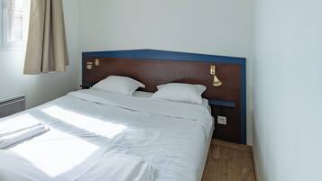 Una cama bien hecha con sábanas blancas, cabecera de madera y una lámpara fijada a la pared.