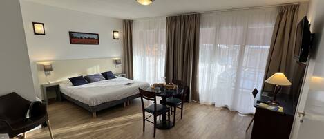 A hotel room with a bed, a desk with a chair, a TV, and a window with curtains.