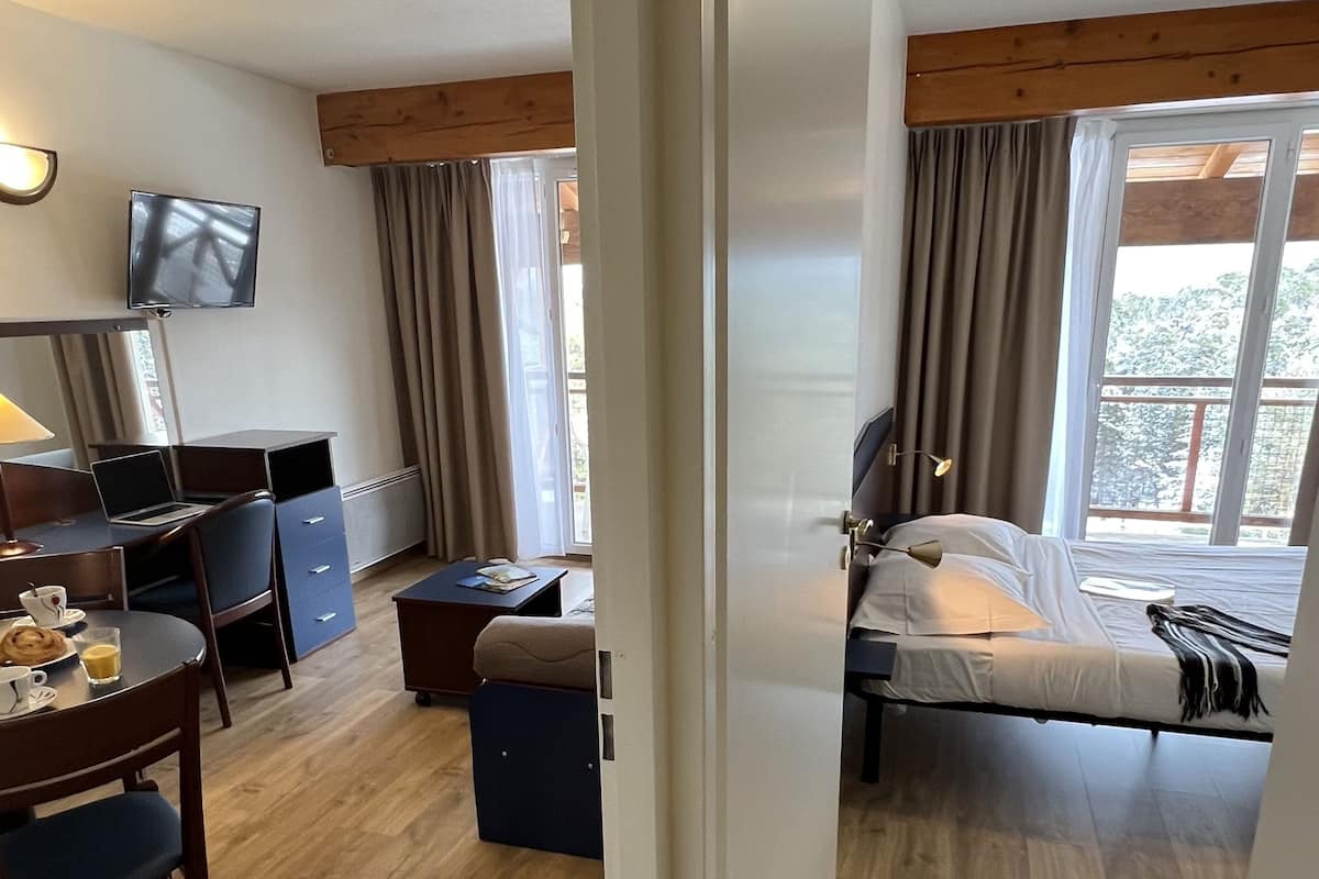 A hotel room with a bed, a desk with a laptop, a chair, a small table with a cup and saucer, and a television on the wall.