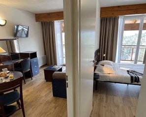 A hotel room with a bed, a desk with a laptop, a chair, a small table with a cup and saucer, and a television on the wall.