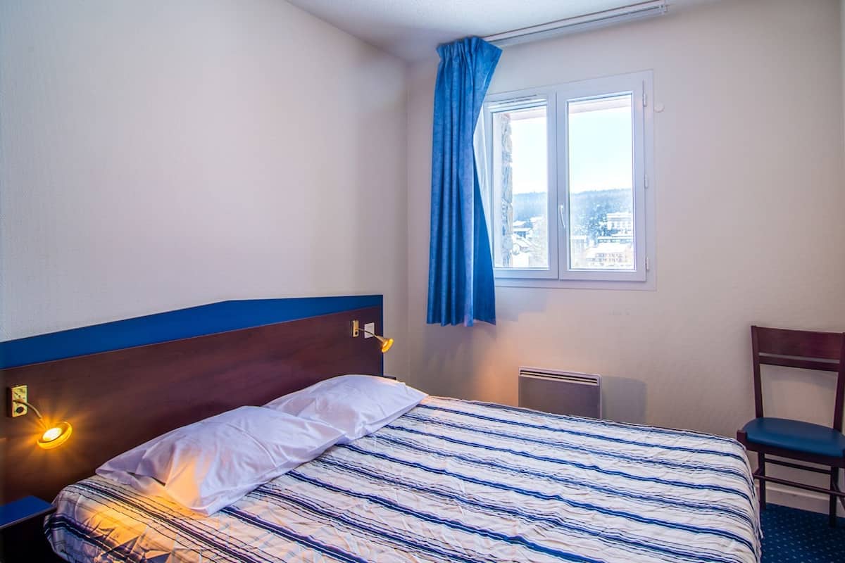 A hotel room with a bed, a chair, a window with curtains, and a wall-mounted lamp.