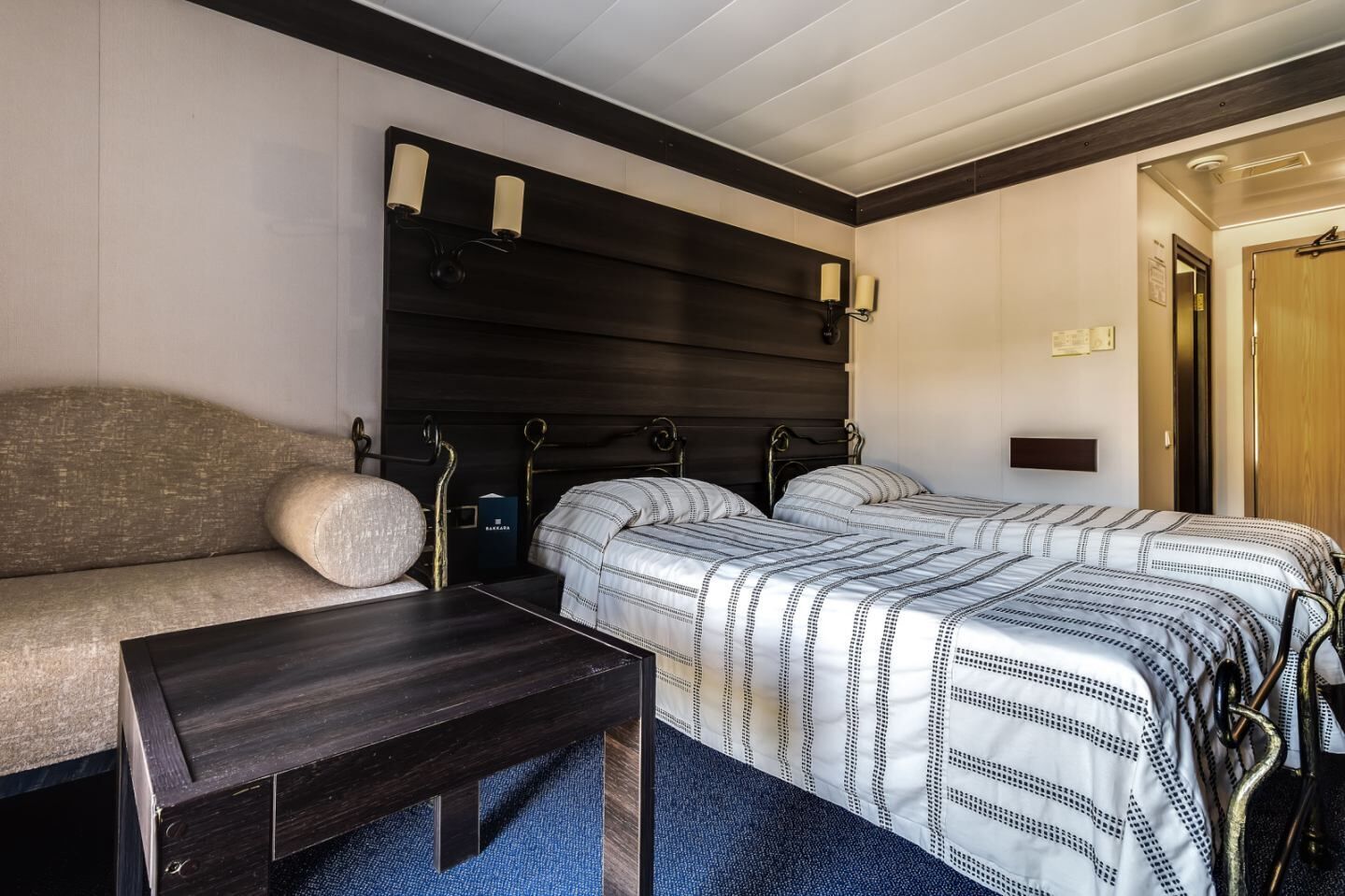 Un dormitorio con cabecera oscura, dos camas con ropa de cama a rayas, una mesita pequeña y un sofá.