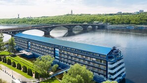 A river cruise ship docked at a pier with a bridge in the background. - Hotel Bakkara (Kyiv)