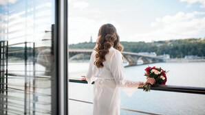 A person in a white dress standing by a window overlooking a river with a bridge, holding a bouquet of flowers. - Hotel Bakkara (Kyiv)