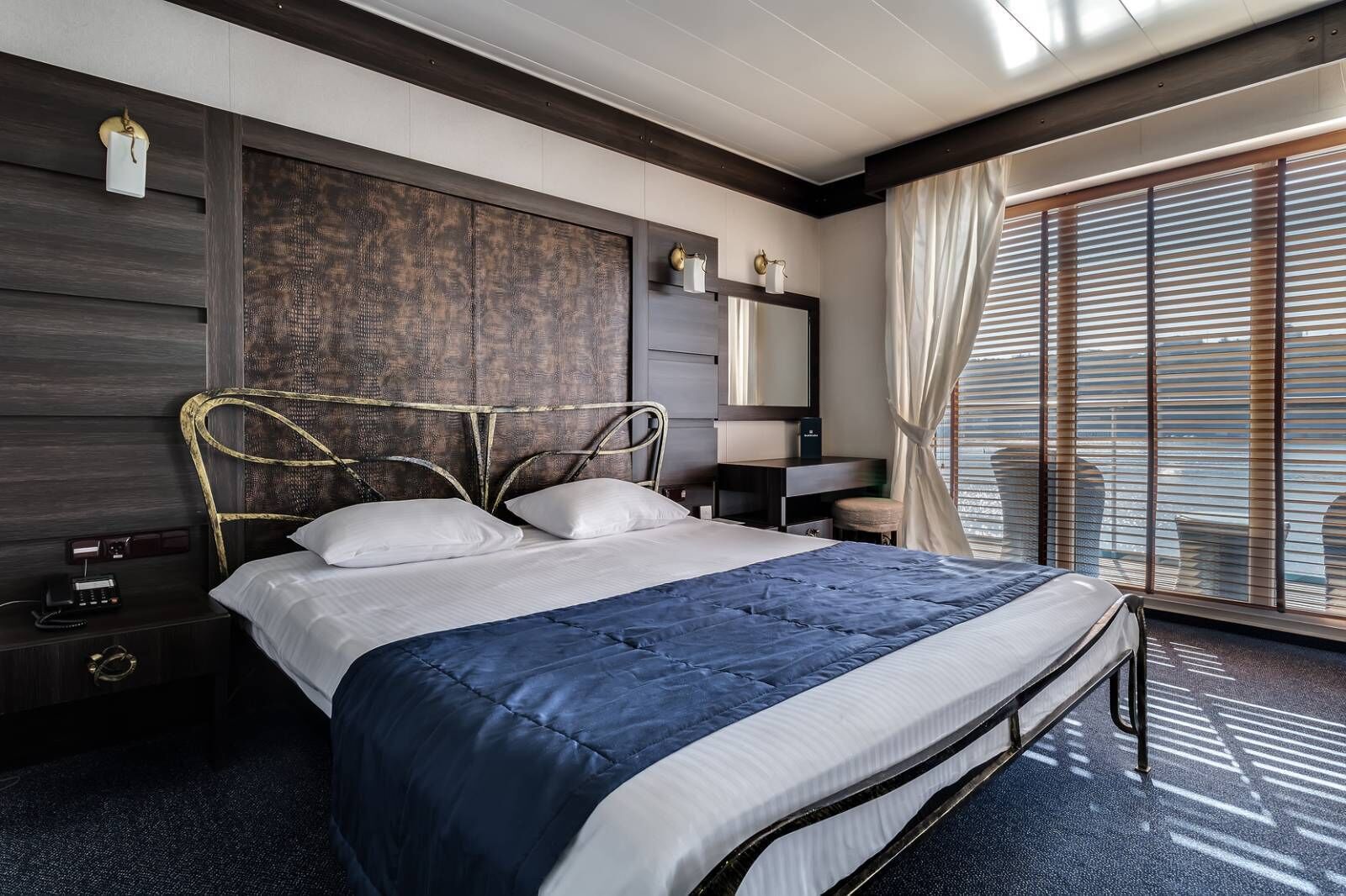 A modern hotel room with a large bed, a bedside table with a lamp, a nightstand, and a large window with blinds.