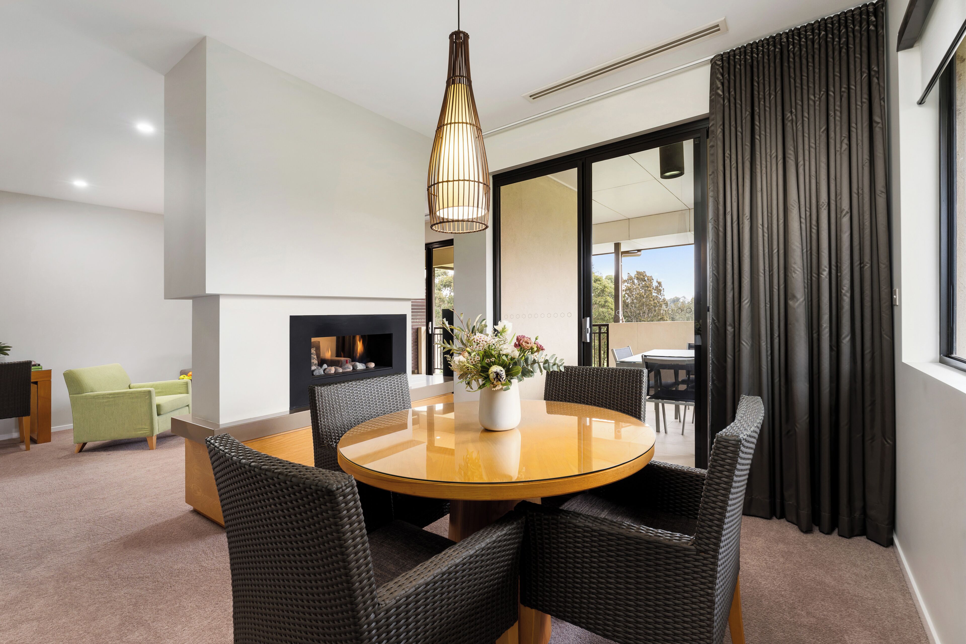 Un salon moderne avec une table ronde en bois, des chaises en osier noir, un luminaire suspendu, une cheminée et une vue dégagée sur l’extérieur grâce à une grande fenêtre ornée de rideaux foncés.