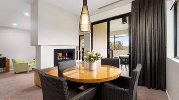 Un salon moderne avec une table ronde en bois, des chaises en osier noir, un luminaire suspendu, une cheminée et une vue dégagée sur l’extérieur grâce à une grande fenêtre ornée de rideaux foncés.