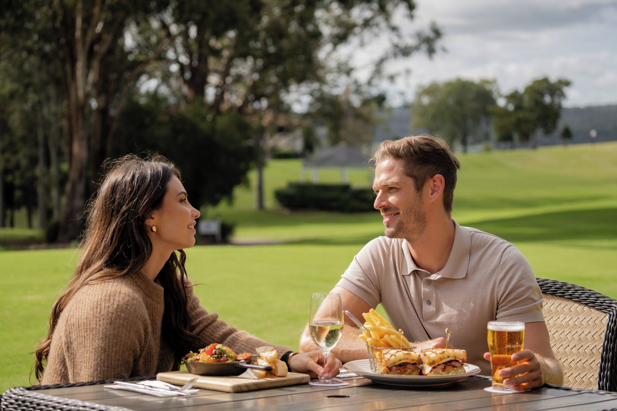 A couple dining outdoors with a table set for two, featuring a meal, a glass of white wine, and a beer.