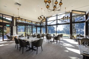 A spacious dining area with multiple tables and chairs, large windows offering a view of the outdoors, and a modern chandelier. - Chateau Elan At The Vintage (Pokolbin)