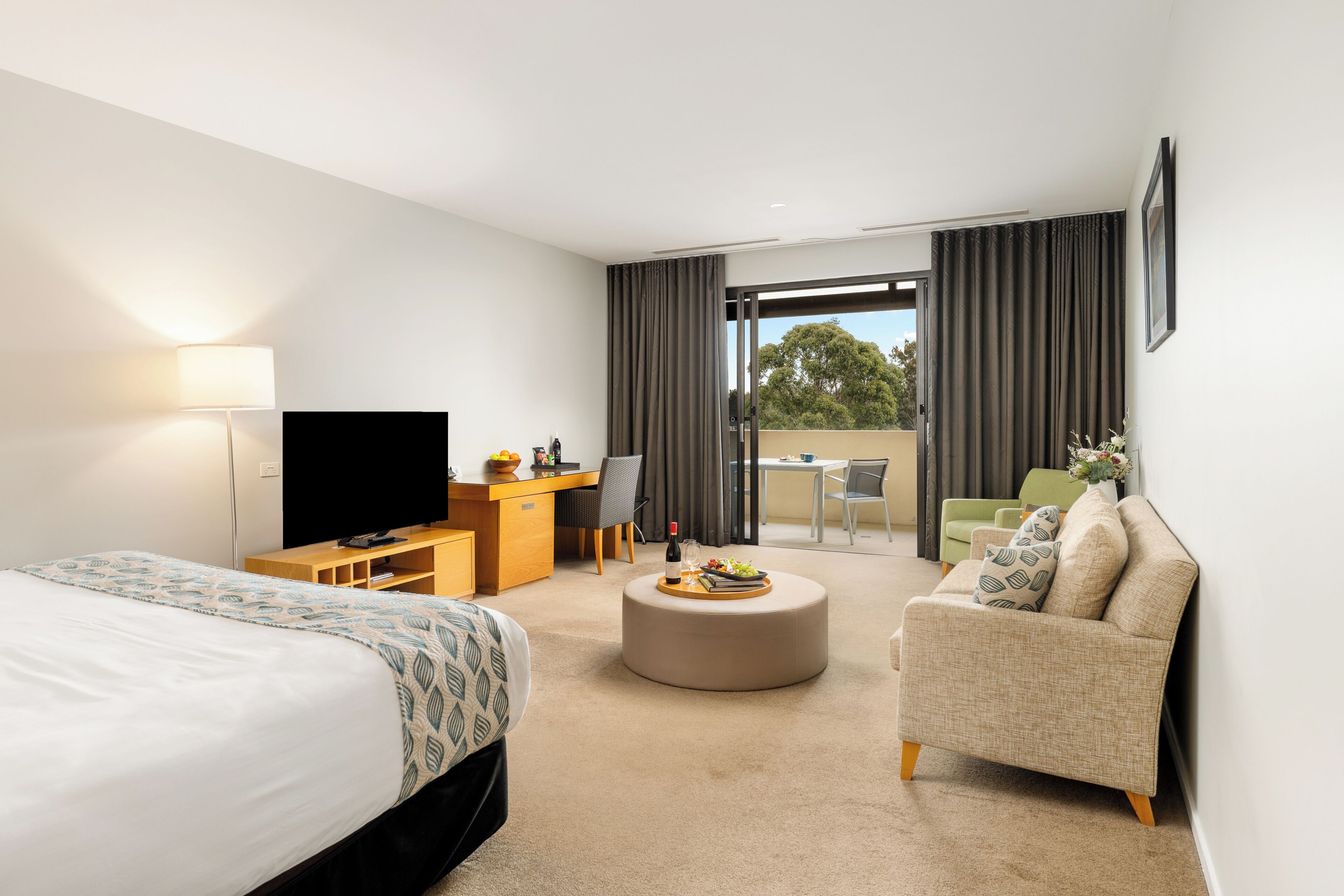 A hotel room with a bed, a desk, a television, a dining area with a table and chairs, and a balcony with curtains.