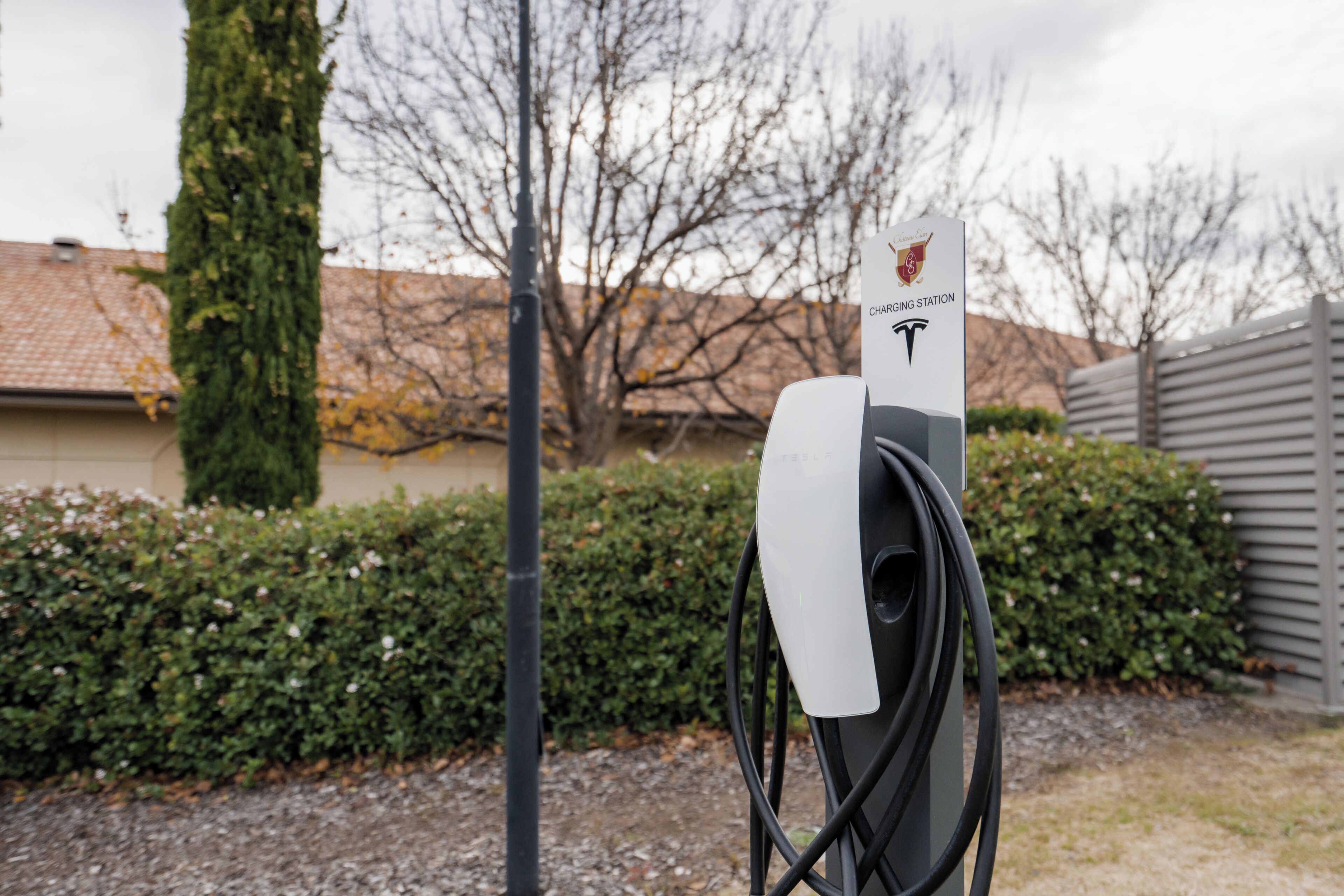 A Tesla charging station with a charging cable connected to it, set against a residential backdrop with a hedge and a house.
