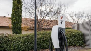 A Tesla charging station with a charging cable connected to it, set against a residential backdrop with a hedge and a house.