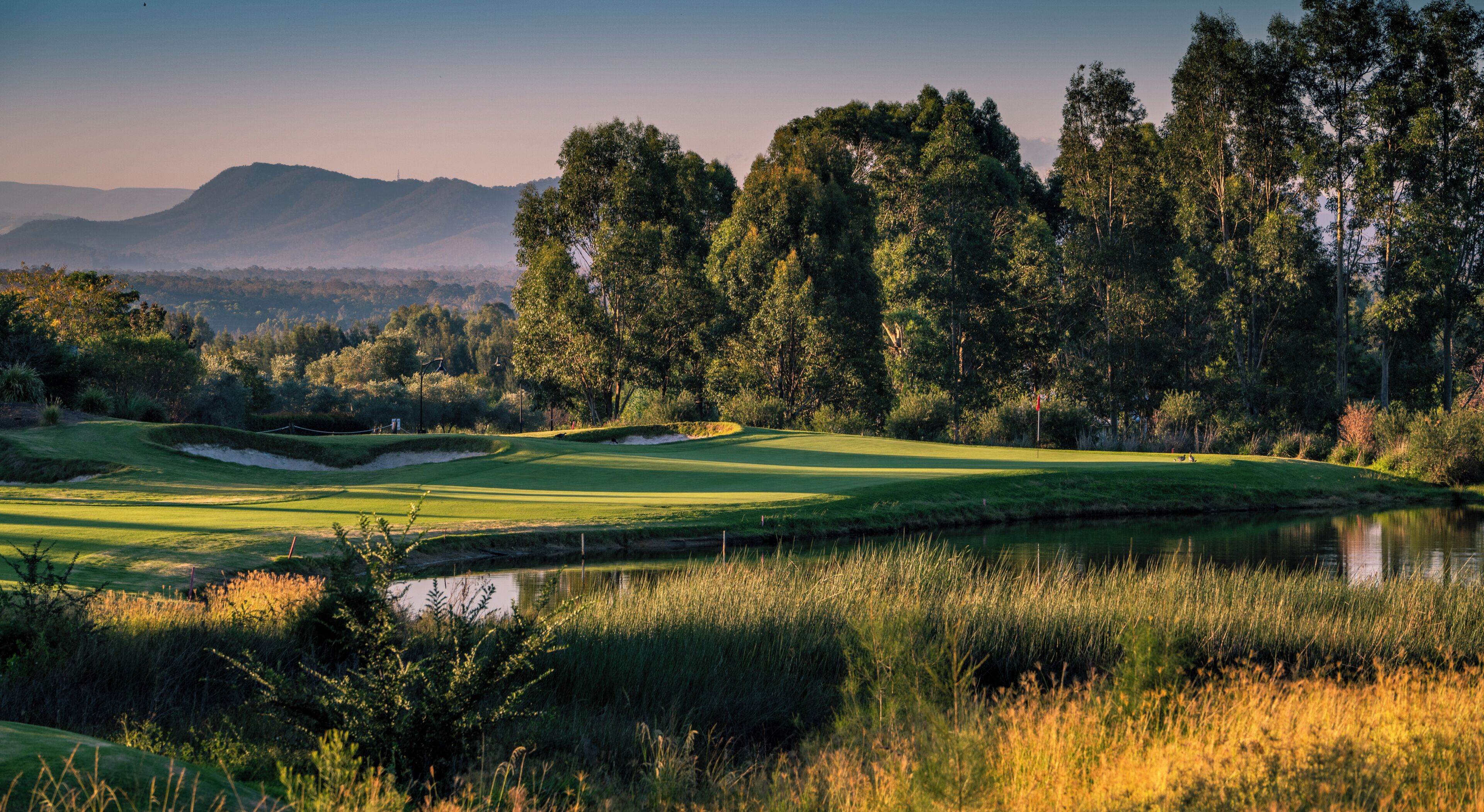 Un campo da golf con uno stagno, alberi e montagne sullo sfondo.
