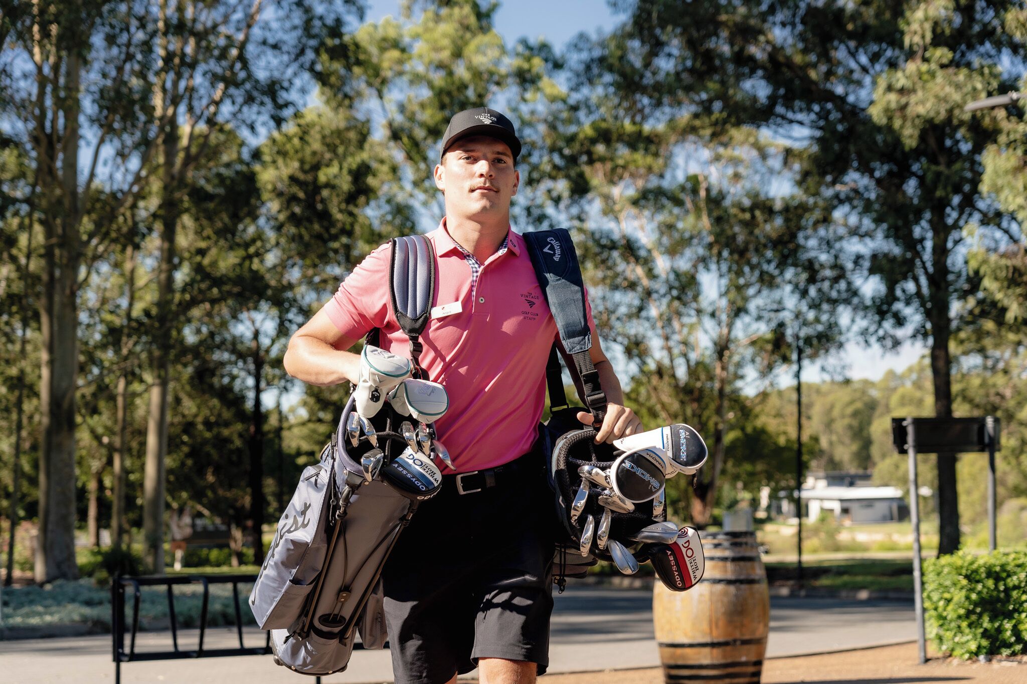 A person carrying golf bags and equipment, walking on a path with trees and a sign in the background.
