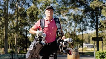 A person carrying golf bags and equipment, walking on a path with trees and a sign in the background.