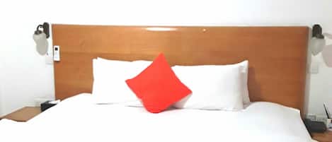 Una cama bien hecha con cabecera de madera, almohada roja y manta blanca.
