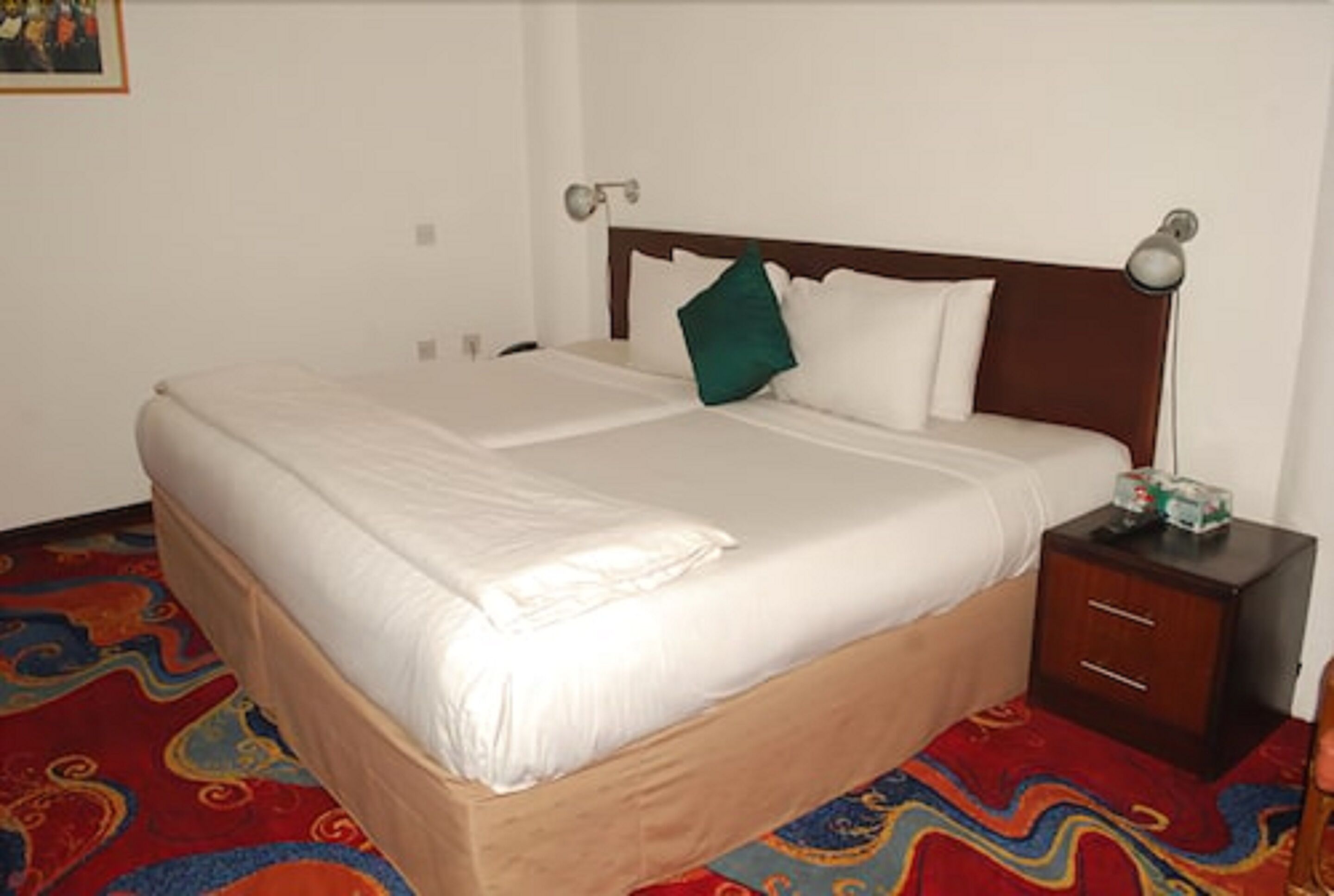 A double bed with white linens, a wooden headboard, a bedside table with a lamp, and a colorful carpet.