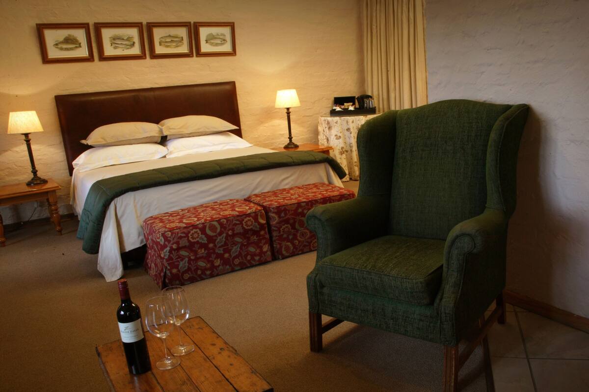 A hotel room with a bed, a green armchair, a wooden table with a bottle of wine and glasses, and a nightstand with a lamp.