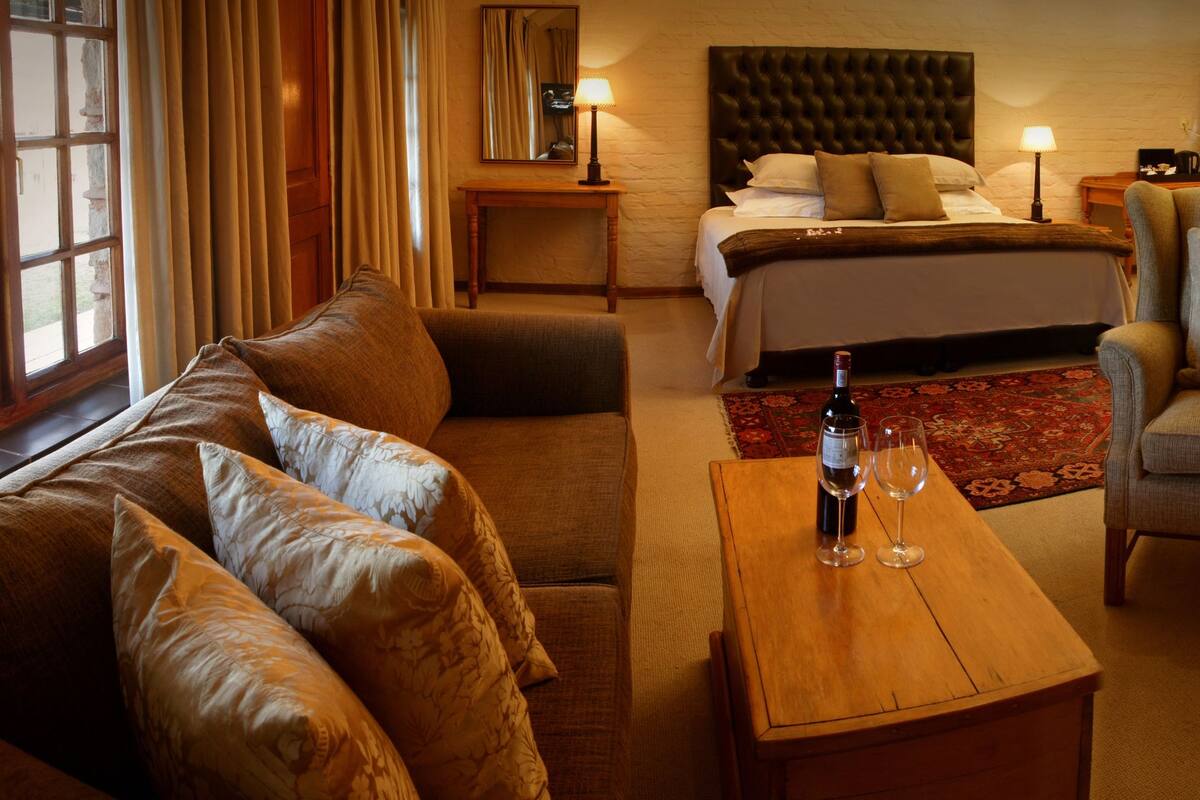 A cozy hotel room with a bed, a sofa, a chair, a small table with a bottle and glasses, a fireplace, and a window with curtains.