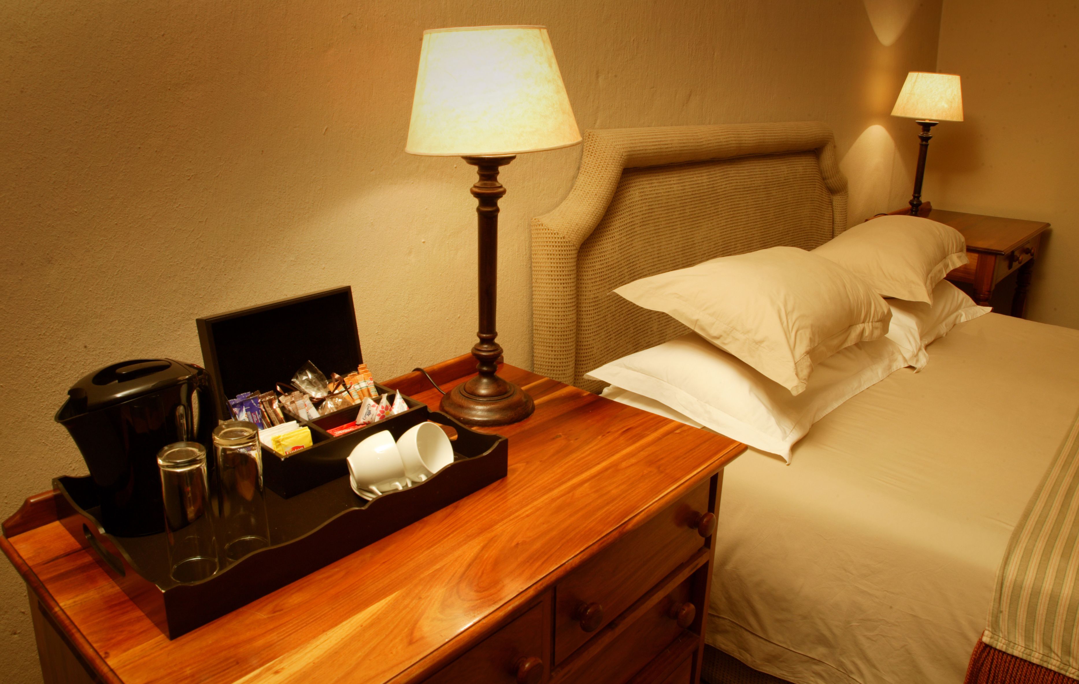 Une chambre d’hôtel avec une table de chevet en bois, un lit avec du linge de maison blanc, deux lampes de chevet, une machine à café et un plateau avec des articles de toilette.