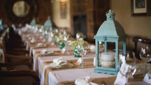 Una larga mesa de comedor con manteles blancos, elegantemente dispuesta con platos, cubiertos y copas de vino. Un farol color turquesa con una vela se coloca sobre la mesa.