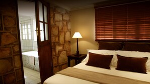 Un dormitorio con pared de piedra, cama con almohadas marrones, mesita de noche con lámpara y ventana con persianas.