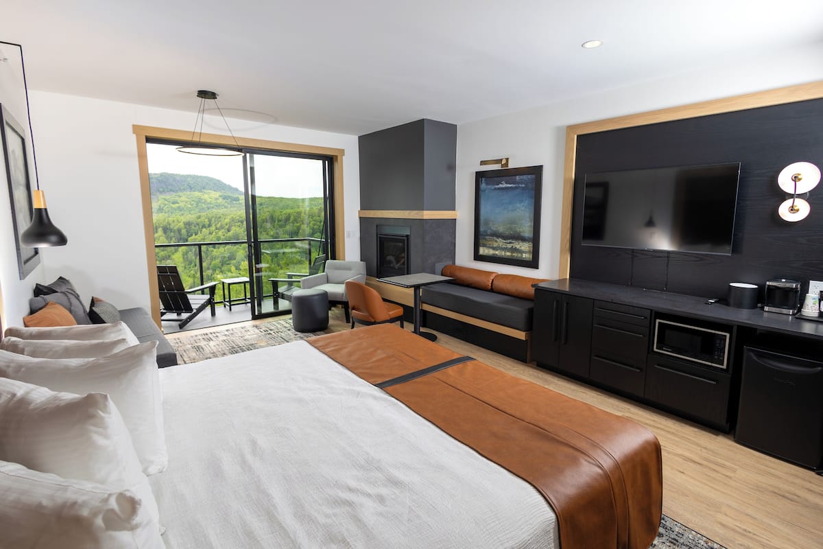 A modern hotel room with a large bed, a sofa, a dining area, a TV, and a fireplace.