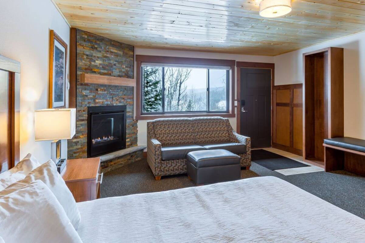 A cozy room with a fireplace, a sofa, a bed, and a large window with a view of trees.