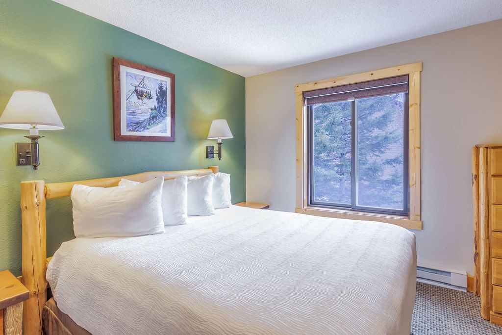 A bedroom with a large bed, two bedside lamps, a framed picture on the wall, and a window with a view of trees.