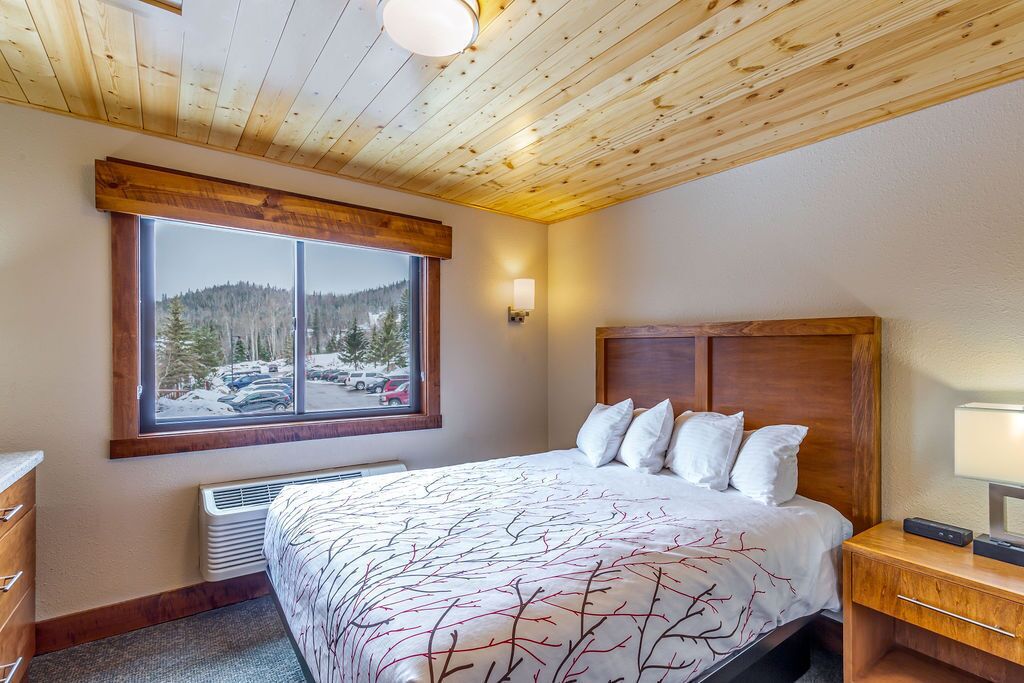 A bedroom with a large bed, a window with a view of a parking lot and trees, a wooden ceiling, and a nightstand with a lamp.