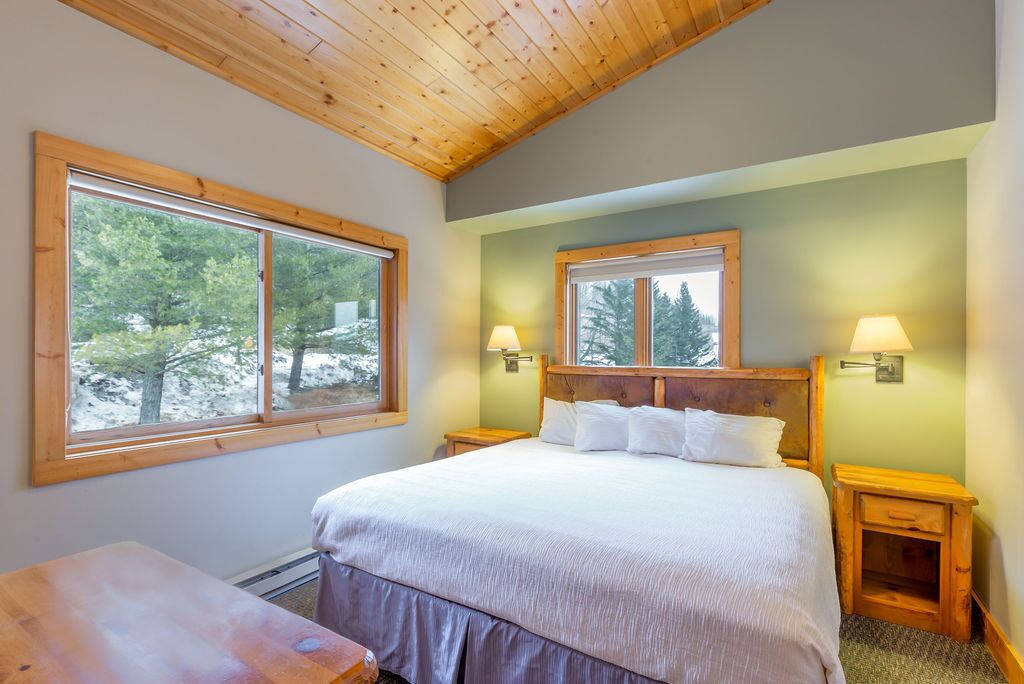 Un dormitorio con una cama grande, dos mesitas de noche, un cabecero de madera y dos ventanas con vistas a árboles.