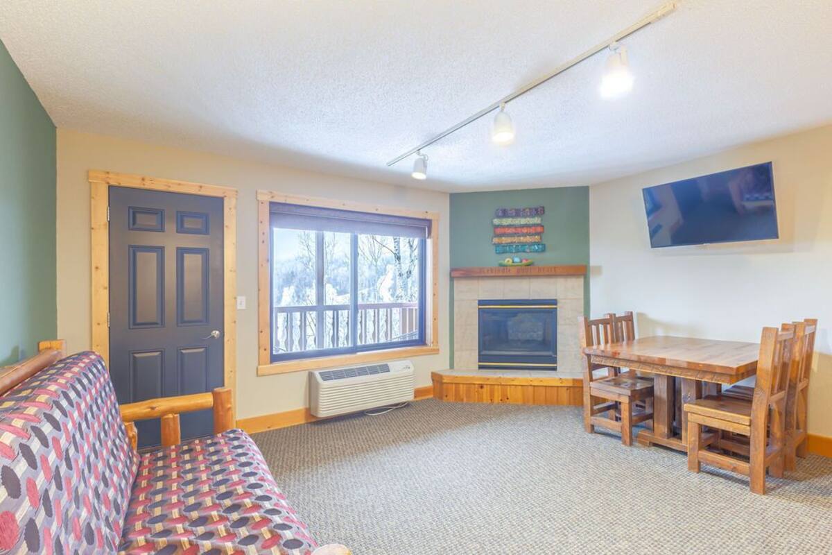 A cozy living room with a sofa, a fireplace, a dining table with chairs, and a large window.