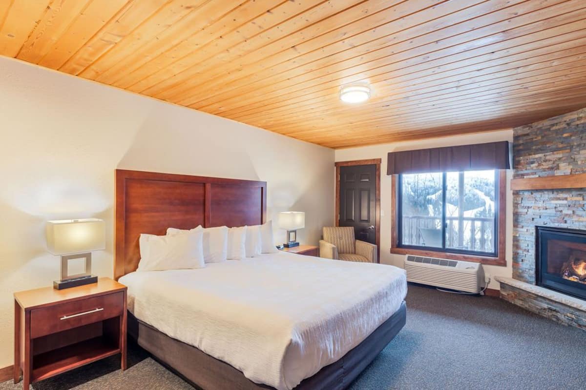 A hotel room with a large bed, wooden headboard, two bedside tables with lamps, a chair, a fireplace, and a stone wall.