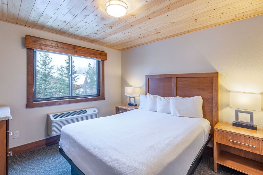 Une chambre d’hôtel avec un grand lit, une tête de lit en bois, des lampes de chevet, une fenêtre donnant sur des arbres et un radiateur intégré.