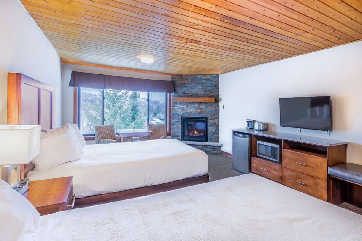A hotel room with two beds, a fireplace, a TV, and a dining area with a view of trees.