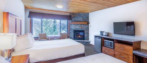 Habitación de hotel con dos camas, chimenea, televisor y zona de comedor con vistas a árboles.