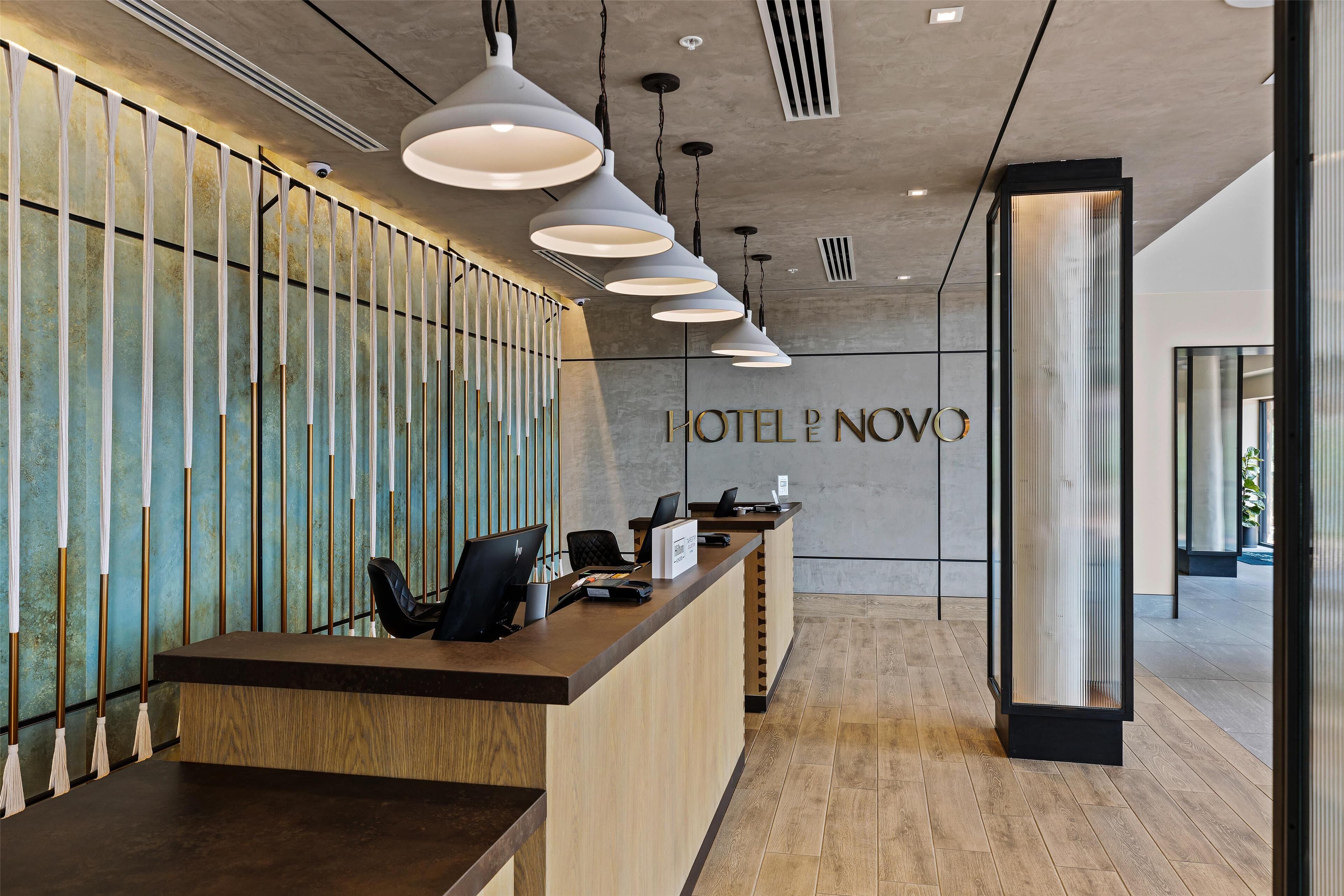 Zona de recepción de un hotel con un mostrador de madera, varias lámparas colgantes y un cartel que dice "HOTEL NOVO".