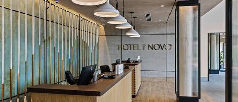 Zona de recepción de un hotel con un mostrador de madera, varias lámparas colgantes y un cartel que dice "HOTEL NOVO".