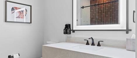 A modern bathroom with a white sink, two faucets, a mirror, and a framed artwork on the wall.