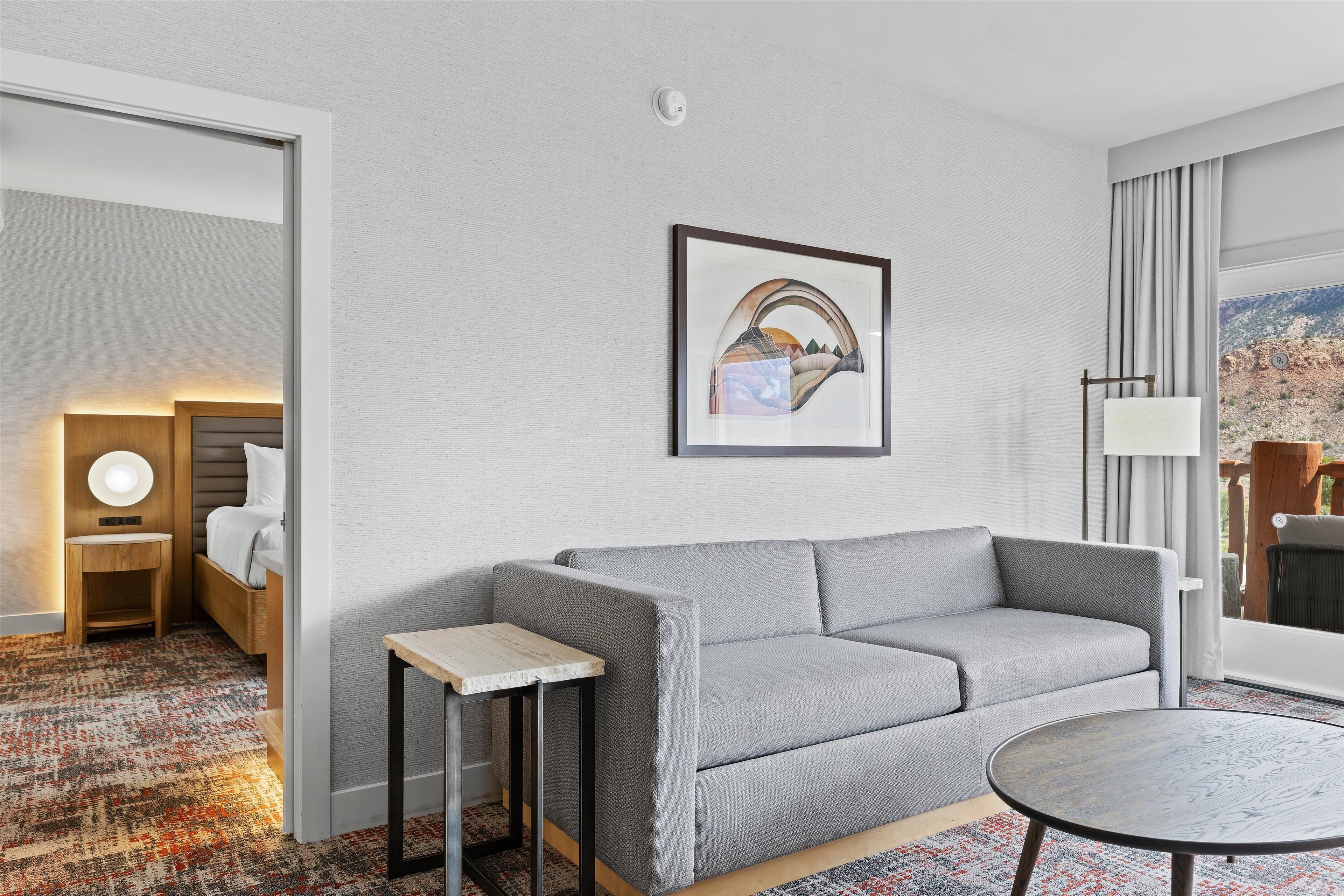 Ein modernes Hotelzimmer mit grauem Sofa, einem kleinen runden Tisch, einem gerahmten Bild an der Wand und Blick ins Freie durch ein Fenster.