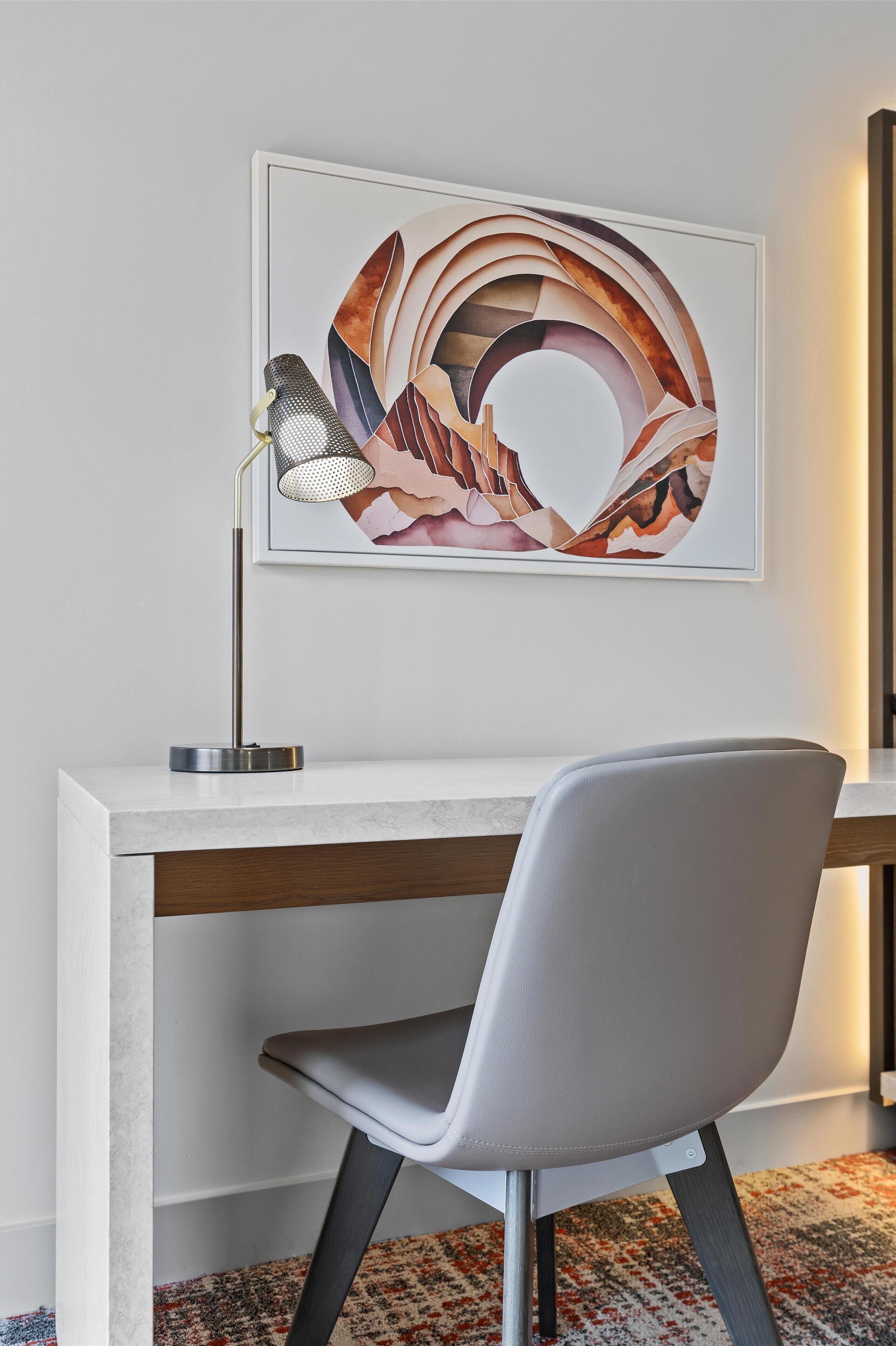 A modern office desk with a chair, a framed abstract painting, and a desk lamp.