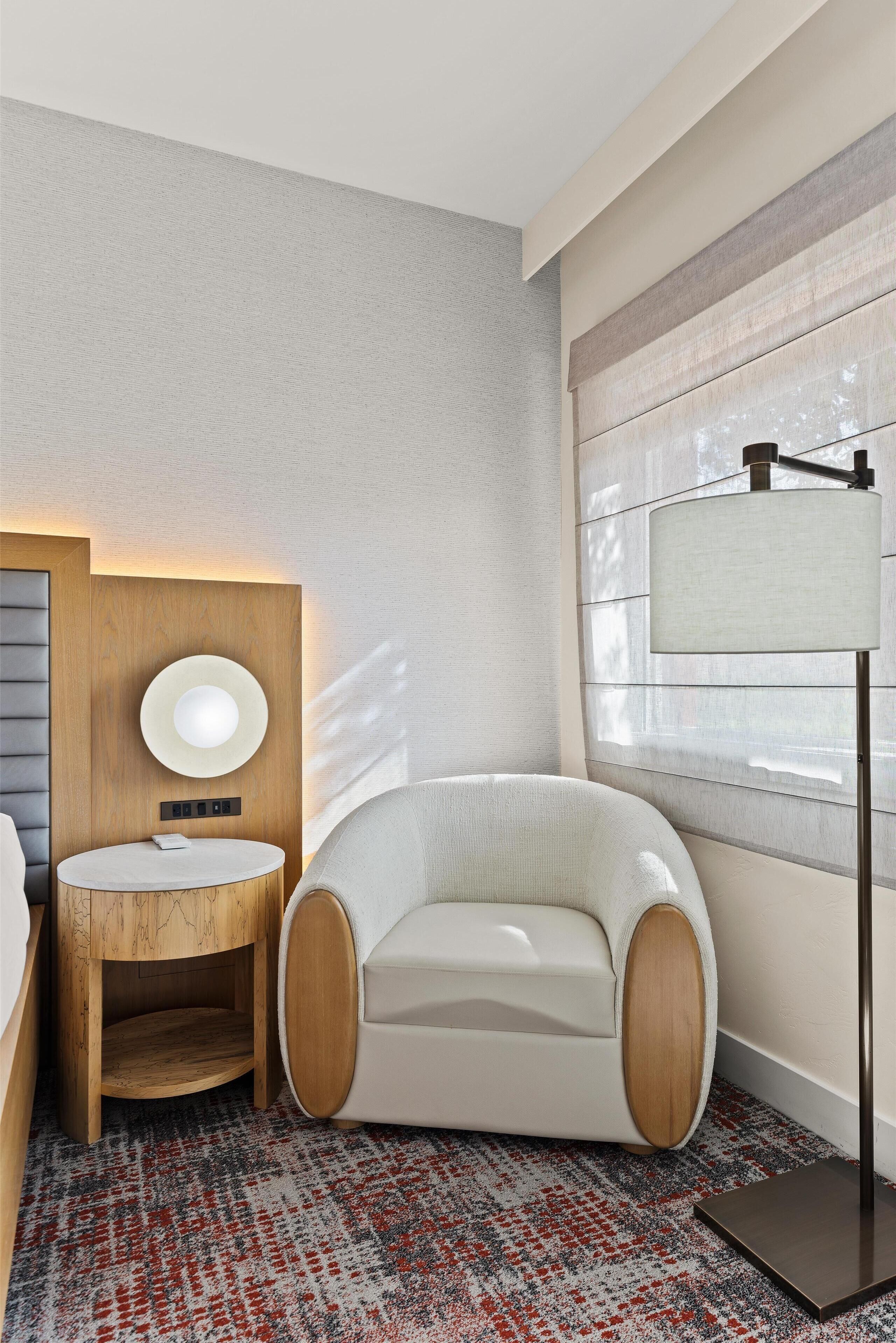 A modern hotel room with a round wooden vanity, a white upholstered armchair, a floor lamp, and a patterned rug.