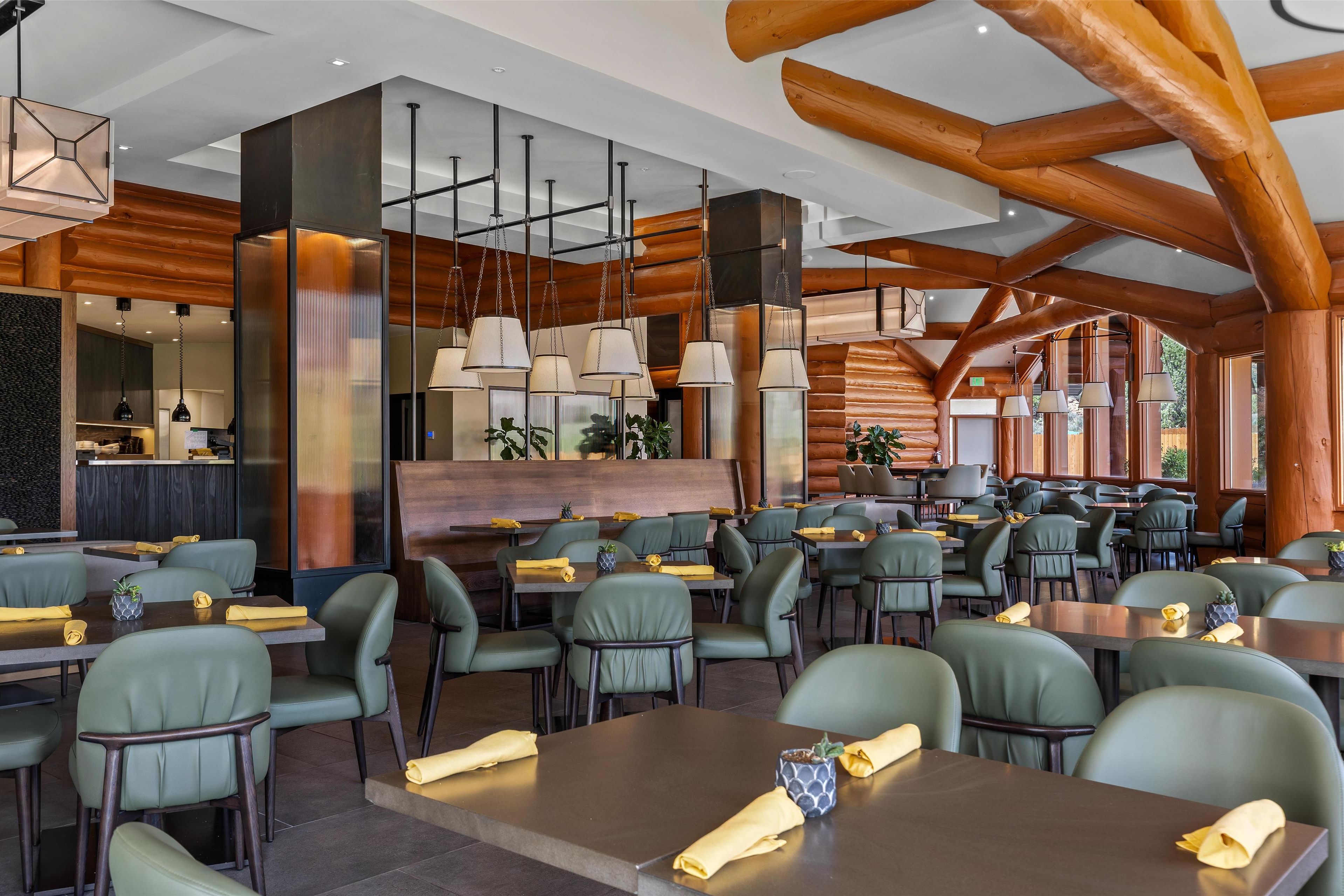 Un restaurant spacieux avec de hauts plafonds, des poutres en bois et un éclairage moderne suspendu. Les tables sont disposées avec soin et agrémentées de chaises à revêtement vert et de serviettes jaunes.